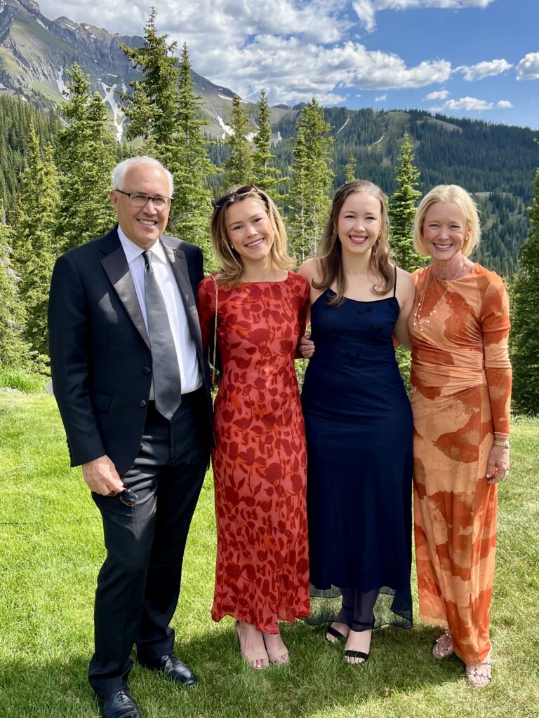 Group of four adults posing outdoors on a grassy slope with pine trees and mountains in the background, dressed in formal attire.