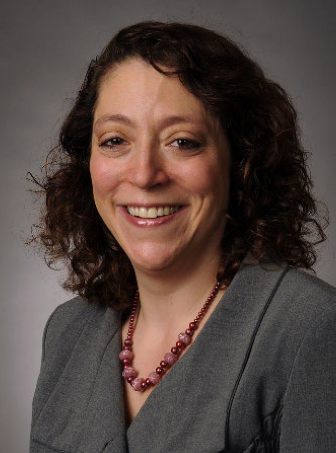 Smiling woman with curly brown hair wearing a gray blazer and pink beaded necklace, facing the camera.