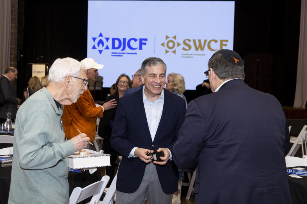 Group of people mingling at a formal event; a smiling man in a navy blazer stands center while others chat around him, with a large logo screen in the background