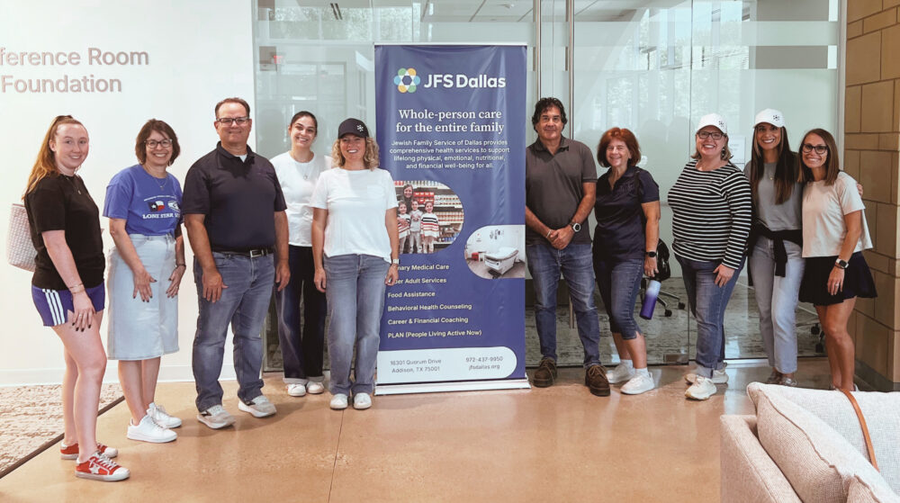 A group of ten adults posing in a lobby beside a blue JFS Dallas banner promoting family care services.