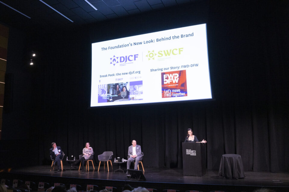 Panel discussion on a dark stage: four guests seated and a speaker at a podium, with a large slide of logos behind them.
