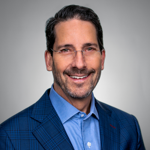 Professional headshot of a smiling man in a blue checkered blazer and light blue shirt, glasses, against a gray backdrop.