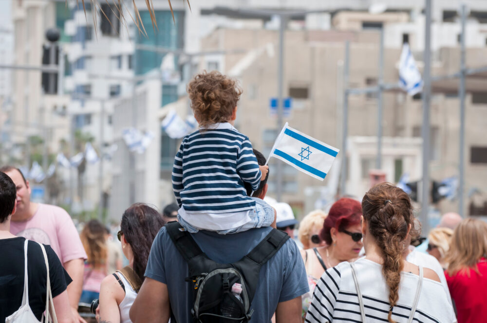 Child sits on an adult's shoulders waving an Israeli flag among a crowded street parade.
