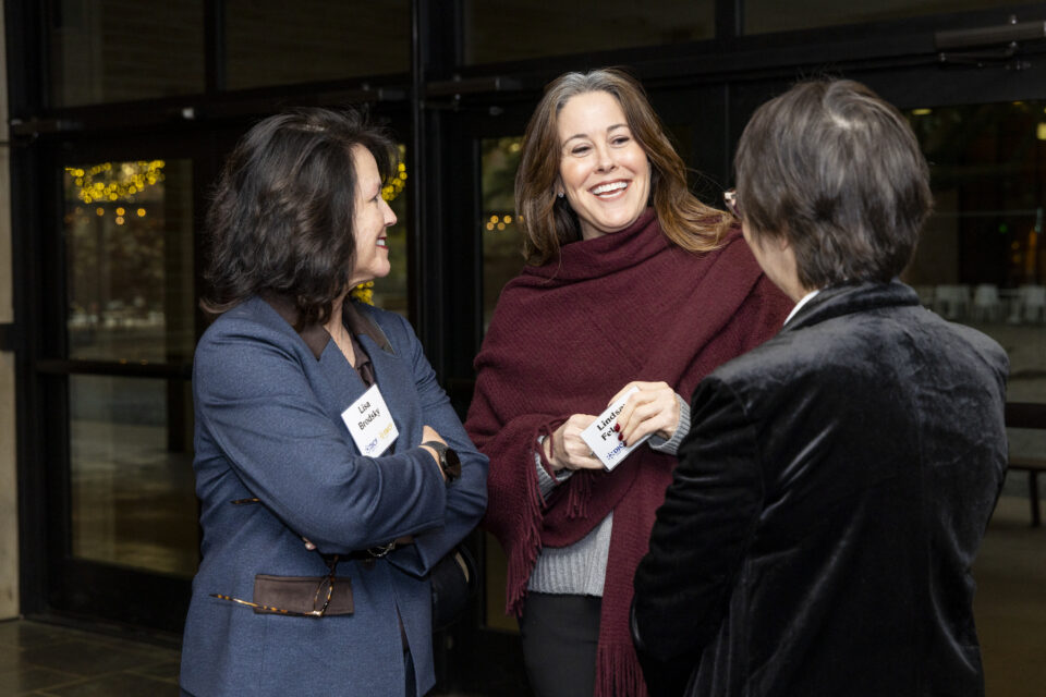 Three women in a hallway, engaged in a friendly conversation, with name badges visible on two of them.
